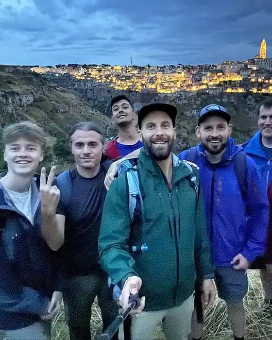 A group of hikers smiling in a selfie at dusk above the gorge, with the lit-up Sassi of Matera behind them