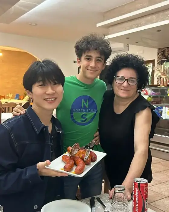 A visitor poses with a Lucanian mother and son at the restaurant table, holding a plate of grilled sausages